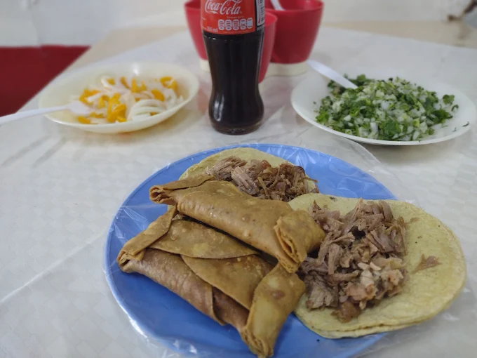 Plato de tacos de carnitas acompañado de salsas, cebolla picada y refresco en un ambiente de restaurante.