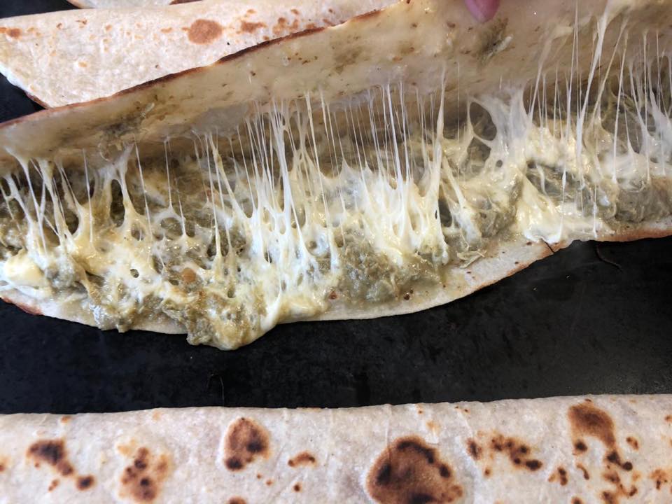 Quesadilla de queso y salsa verde, con queso fundido estirándose, presentada sobre tortillas en una superficie oscura.