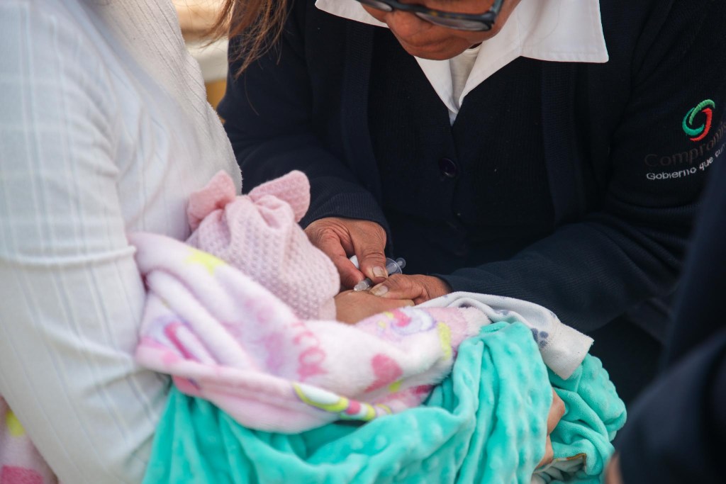 Una trabajadora de salud está administrando una vacuna a un bebé envuelto en mantas mientras es sostenido por un adulto en un evento de vacunación.