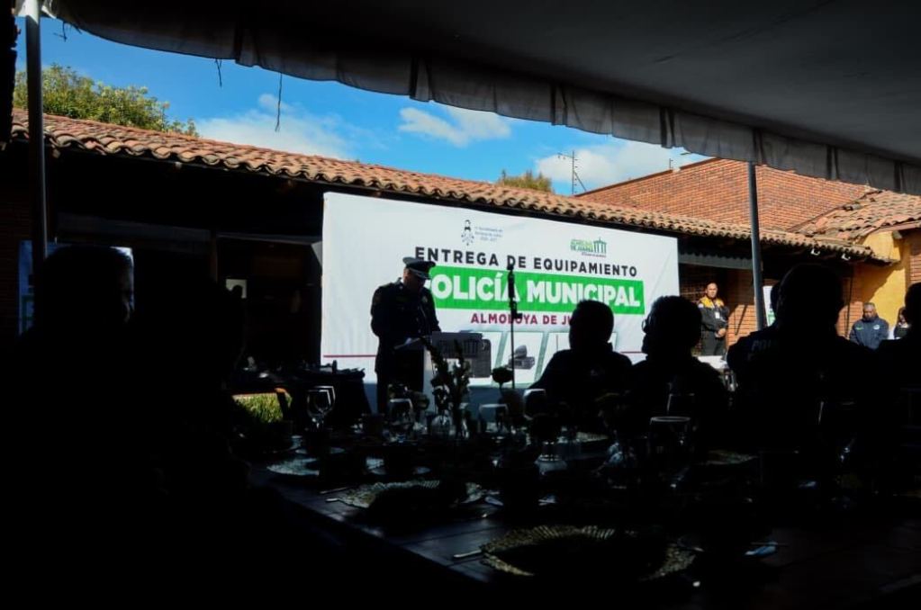 Evento de entrega de equipo policial con el alcalde hablando frente a un cartel que dice 'Entrega de Equipamiento Policía Municipal Almoloya de Juárez' en un ambiente al aire libre.