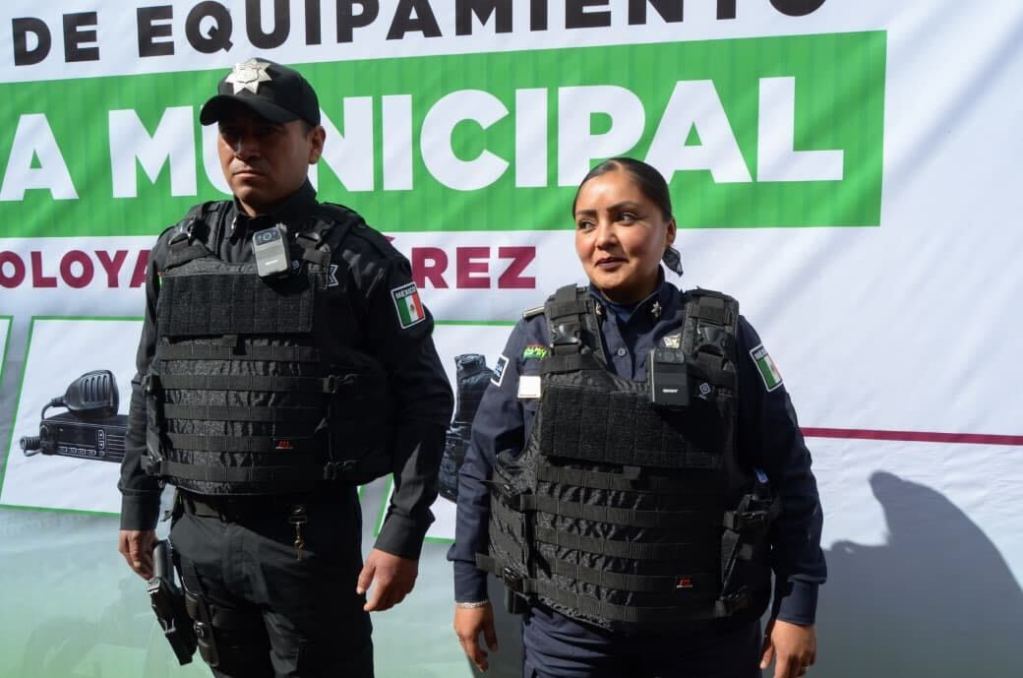 Dos policías con chalecos antibalas posan frente a un banner que destaca la entrega de equipo para la Policía Municipal de Almoloya de Juárez.