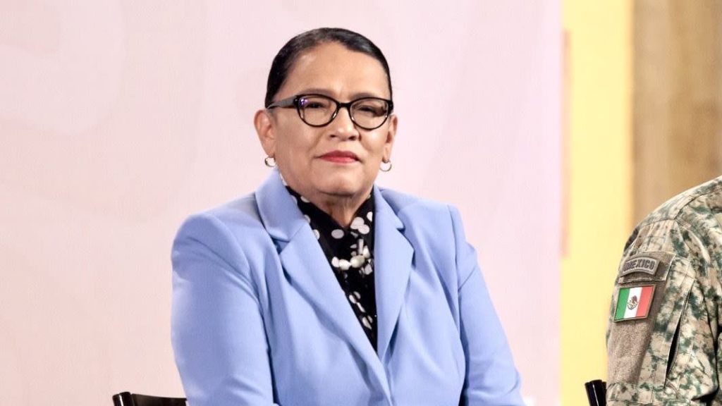 Una mujer con gafas y un blazer azul, sentada frente a un fondo neutro, observa de manera seria.