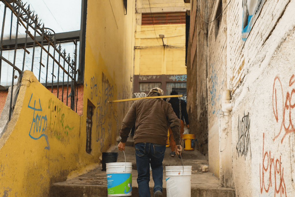 Hombre cargando cubetas de agua mientras camina por una calle estrecha con paredes graffiti.