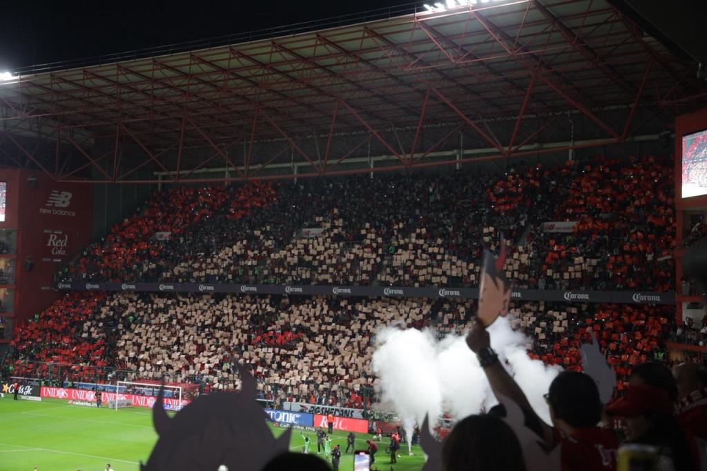 Aficionados en el estadio Nemesio Diez creando un tifo colorido para un partido de fútbol, con banderas rojas y blancas y humo en el aire.