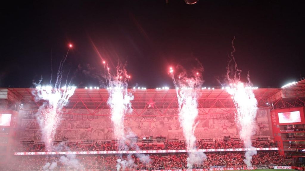 Fuegos artificiales iluminando el estadio Nemesio Diez durante un partido de fútbol, con un gran mural de fondo y una multitud de aficionados animando.