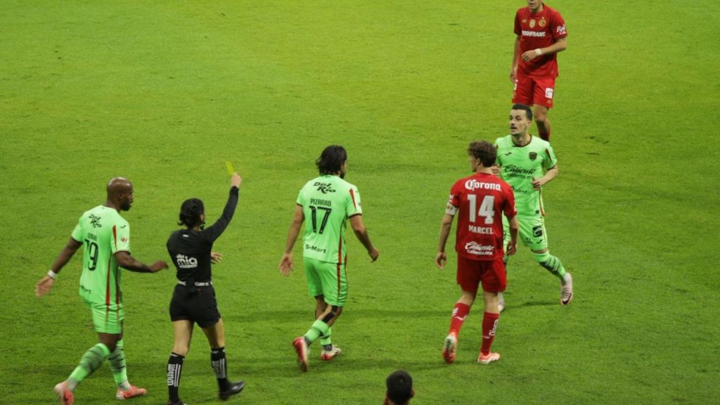 Jugadores en el campo de fútbol, uno de ellos recibiendo una tarjeta amarilla de la árbitra, mientras otros se comunican entre ellos.