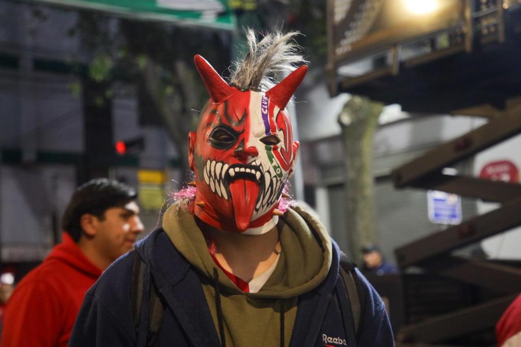 Hombre con máscara demoníaca y cuernos, en un ambiente de partido de fútbol.