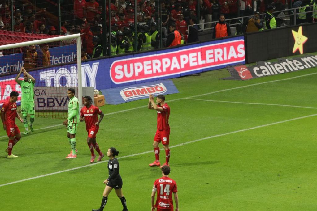Jugadores del equipo de Toluca en acción durante un partido de futbol, con un portero rival visible detrás. El ambiente es tenso y emocionante, con los aficionados en las gradas animando.
