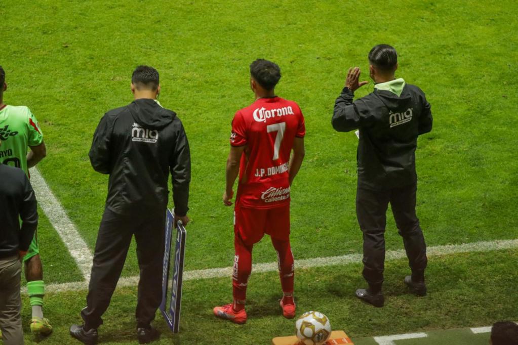 Jugador de fútbol en la banca, esperando ser sustituido durante un partido. Lleva una camiseta roja con el número 7 y el nombre 'Dominguez'. Entrenador y personal técnico a su lado.