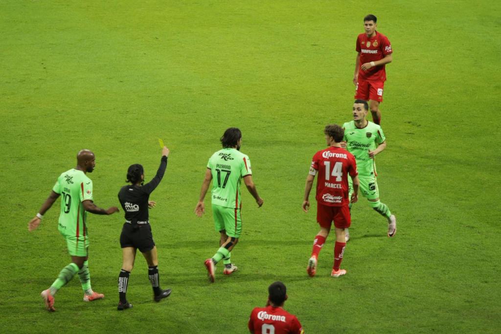 Un árbitro mostrando una tarjeta amarilla a un jugador del equipo con uniforme verde durante un partido de fútbol, con otros jugadores y un asistente en el fondo.