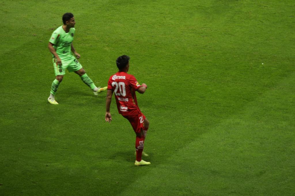 Jugadores en el campo durante un partido de fútbol, con uno vestido de rojo y el otro de verde.