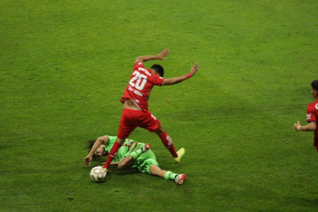Jugador con camiseta roja número 20 salta sobre un jugador del equipo contrario caído en el campo de fútbol, mientras otros jugadores observan la acción.
