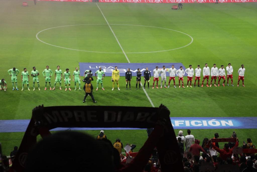 Equipos alineados en el campo de futbol antes del inicio del partido entre Bravos de Juárez y Toluca, con la afición sosteniendo una bufanda en primer plano.