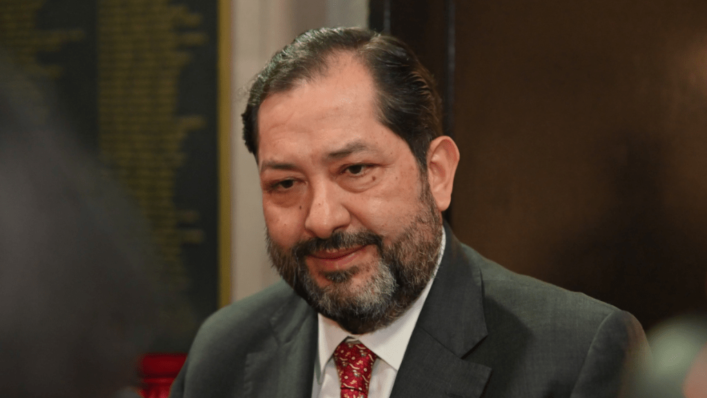 Retrato de un hombre con barba y traje, sonriendo levemente, en un entorno interno con paredes oscuras.