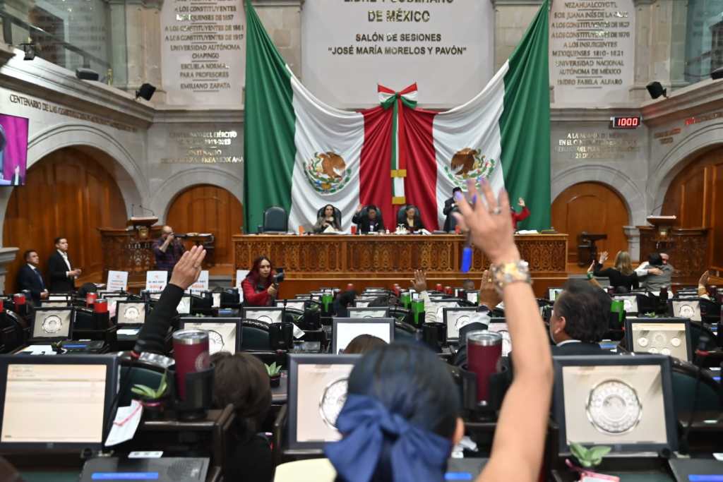 Vista del salón de sesiones del Congreso del Estado de México, con diputadas y diputados levantando la mano en señal de votar. Al fondo, se destaca un gran retrato de la bandera mexicana y el presídium.