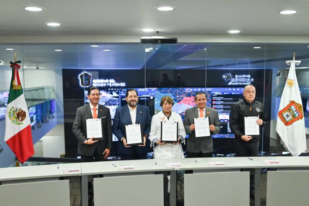 Funcionarios de diferentes instituciones sosteniendo documentos en una ceremonia, con banderas de México en ambos lados y pantallas de monitoreo al fondo.
