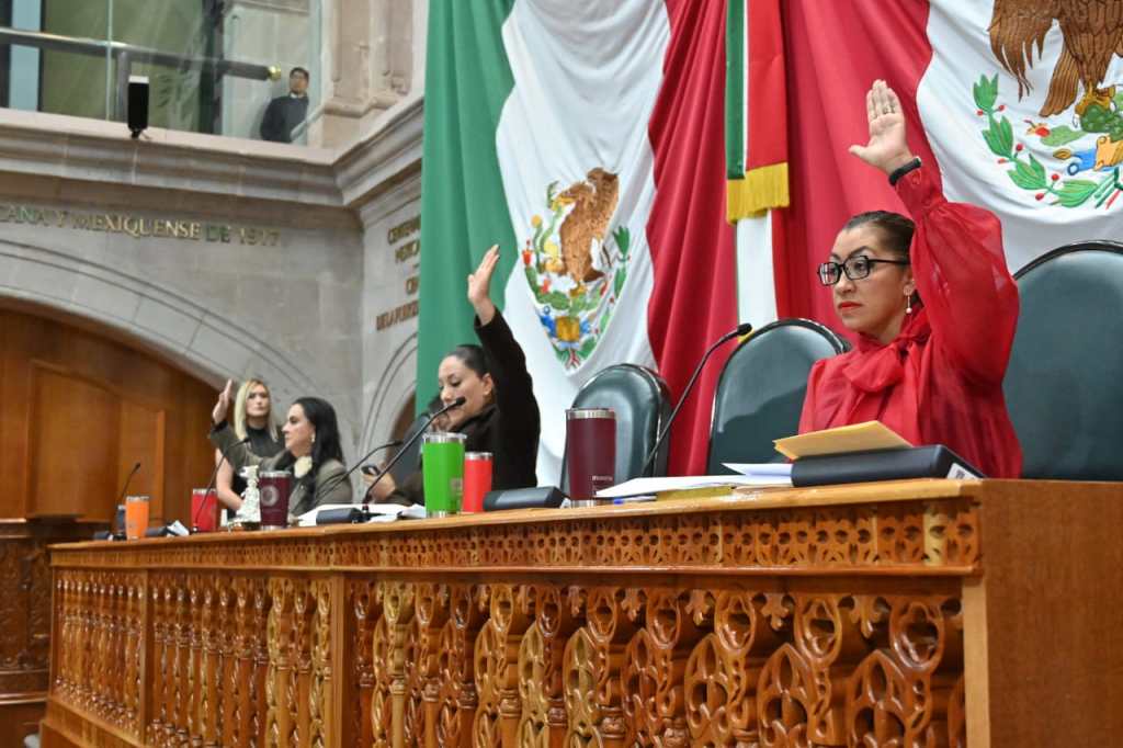 Diputadas en el Congreso del Estado de México levantando la mano durante una sesión, con banderas de México y el escudo nacional de fondo.