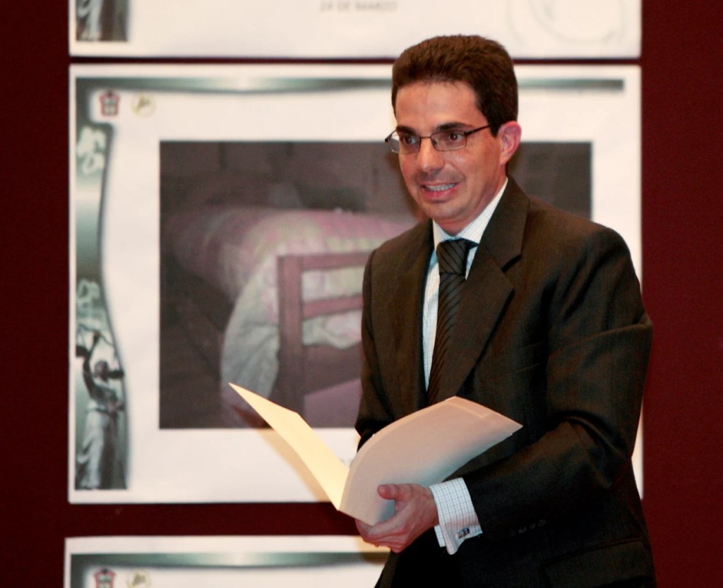 Alberto Bazbaz presentando documentos en un evento, con una imagen de una cama visible en el fondo.