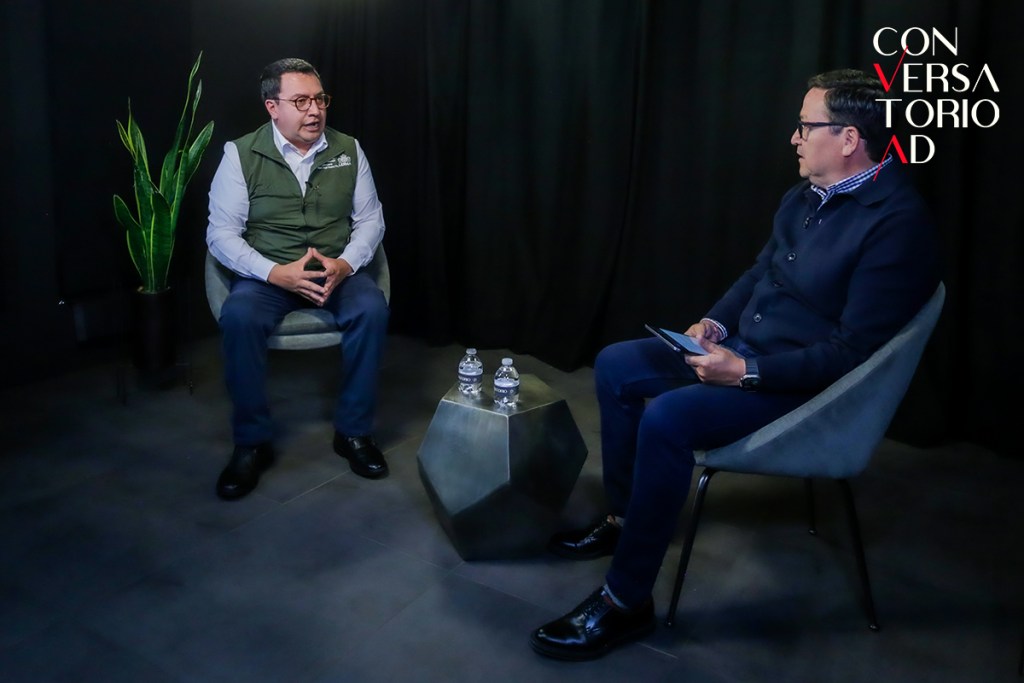 El alcalde de Lerma, Miguel Ángel Ramírez Ponce, y el periodista Mario García Huicochea durante una entrevista en un ambiente formal, sentados en sillas frente a un decorado minimalista con una planta de interior.