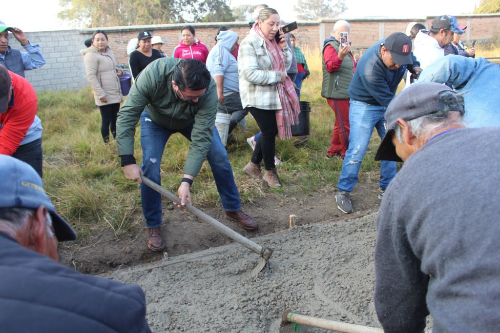 El presidente municipal Adolfo Solís Gómez participa en la construcción de una obra comunitaria con la ayuda de vecinos, utilizando herramientas de construcción en un terreno al aire libre.