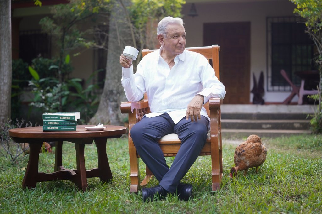 Andrés Manuel López Obrador sentado en una silla, sosteniendo una taza en un entorno verde y natural, con un libro de fondo en una mesa y una gallina en el suelo cerca de él.