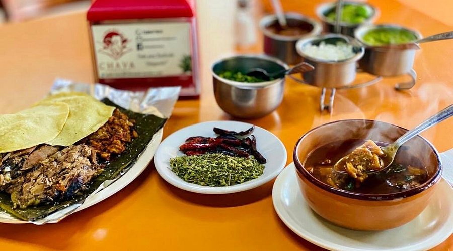 Plato de barbacoa con tortillas, consomé humeante y acompañamientos como chiles secos y hierbas, presentado sobre una mesa de color naranja.