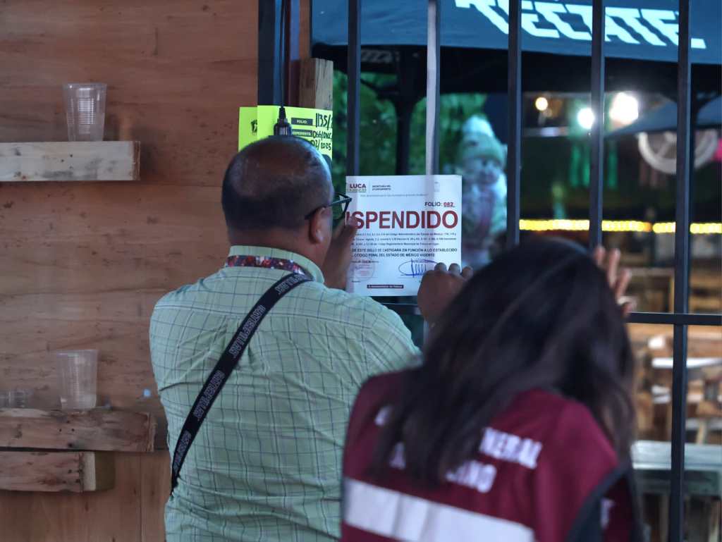 Un funcionario coloca un aviso de clausura en la entrada de un bar, mientras otro individuo observa. Se pueden ver mesas y sillas vacías dentro del establecimiento.
