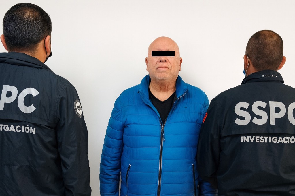 Hombre de pie vestido con chaqueta azul mientras es supervisado por dos agentes de la ley uniformados.