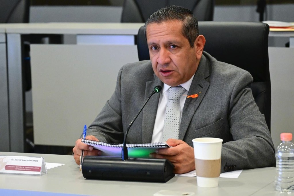 El magistrado Héctor Macedo García durante una reunión, sosteniendo un cuaderno y hablando al micrófono, con una taza de café en la mesa.