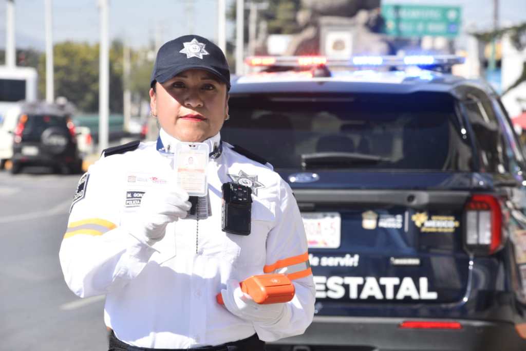 Agente de tránsito mostrando su identificación y terminal electrónica portátil en una vía pública, con un vehículo de policía de fondo.