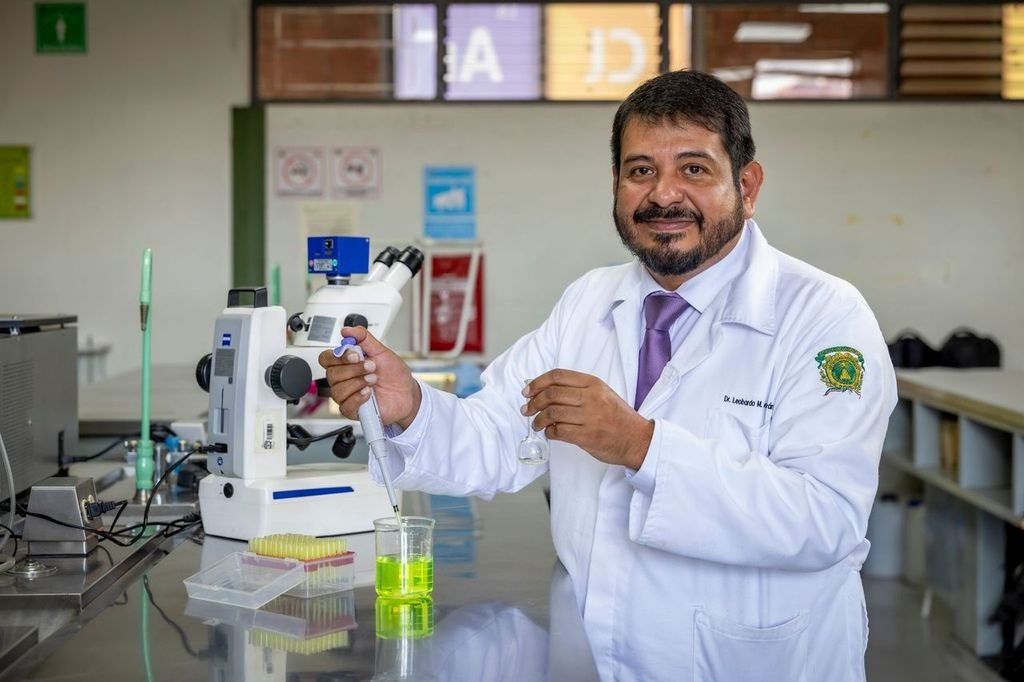 El profesor Leobardo Manuel Gómez Oliván sosteniendo un vaso de precipitados con líquido verde y una micropipeta en un laboratorio, con un microscopio y equipo de laboratorio al fondo.