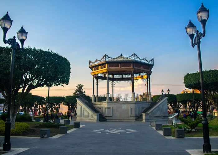 Kiosk en un parque al atardecer, rodeado de árboles y faroles, con una vista despejada del cielo.