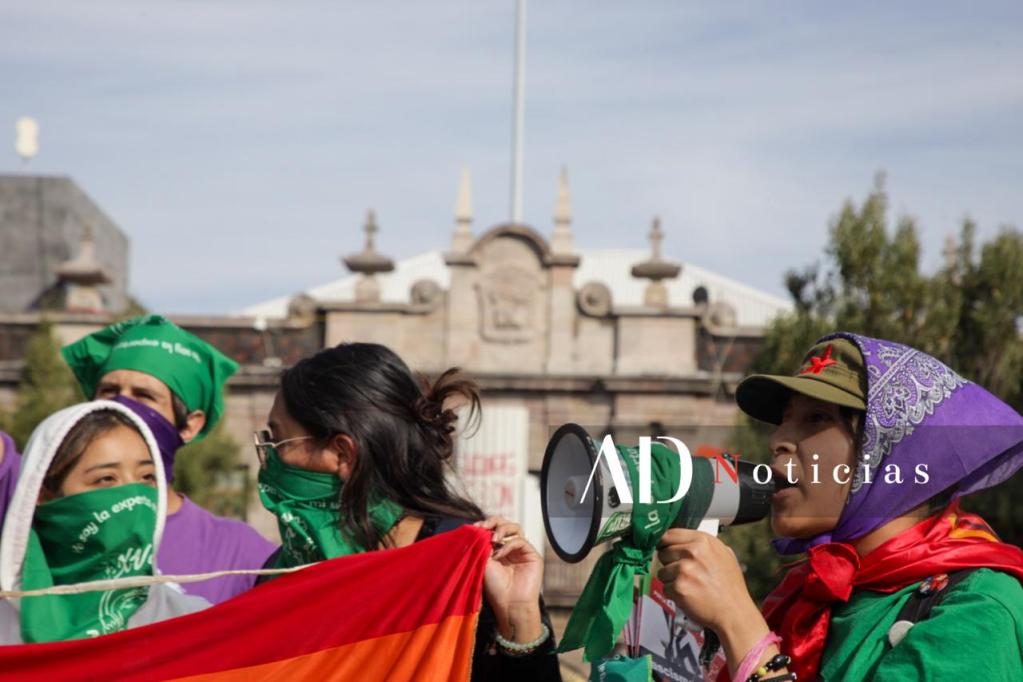 Grupo de feministas en una manifestación en Toluca, sosteniendo una bandera multicolor y utilizando pañuelos verdes, mientras una de ellas habla por un megáfono.
