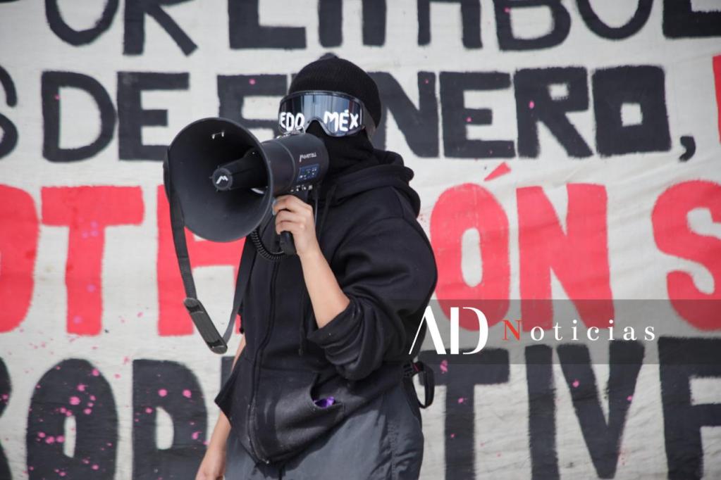 Una activista feminista sostiene un megáfono durante una protesta, con un fondo que incluye un cartel relacionado con la violencia de género en el Estado de México.