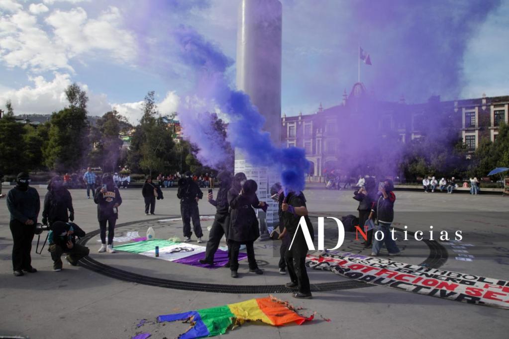 Activistas feministas en Toluca utilizan humo de color morado durante una manifestación con motivo del 25 de noviembre, Día Internacional de la Eliminación de la Violencia contra la Mujer. Se observan pancartas y una bandera con los colores LGBTTIQ+ en el suelo.