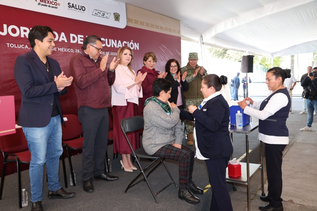 Evento de vacunación en Toluca, donde se observa a un grupo de personas, incluyendo autoridades, aplaudiendo mientras una mujer recibe una vacuna por parte de personal de salud.