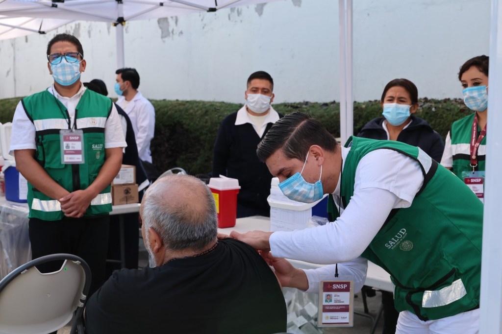 Healthcare worker administering a vaccine to a person seated, with several medical staff observing in a vaccination setting.