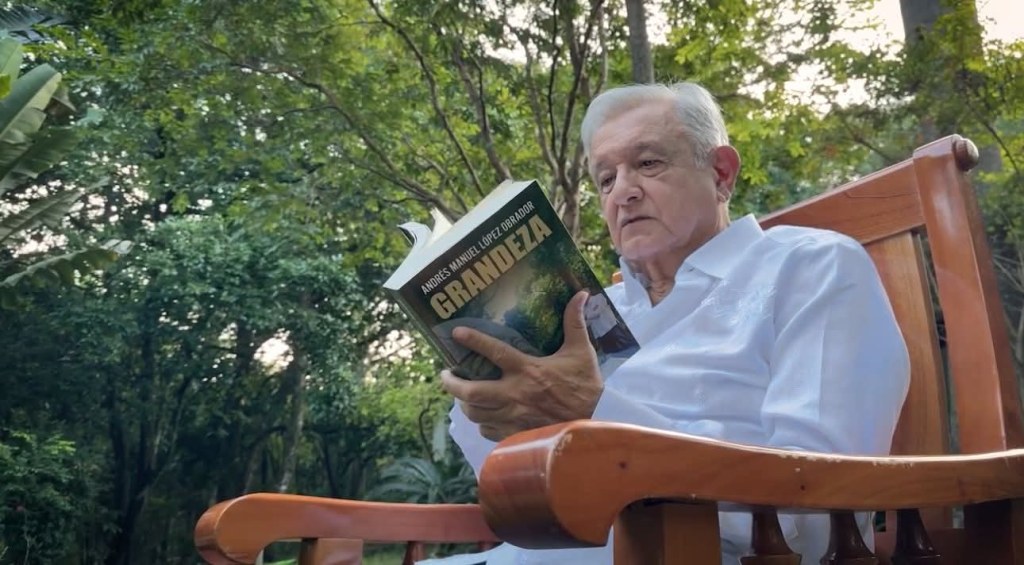 Andrés Manuel López Obrador sentado en una silla de madera, leyendo su libro 'Grandeza' en un entorno natural con árboles y vegetación de fondo.