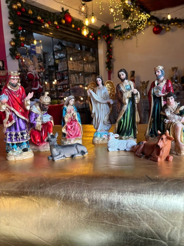 Figuras de un nacimiento navideño con personajes como María, José, los Reyes Magos y Ángeles, sobre una mesa decorada con un fondo festivo.