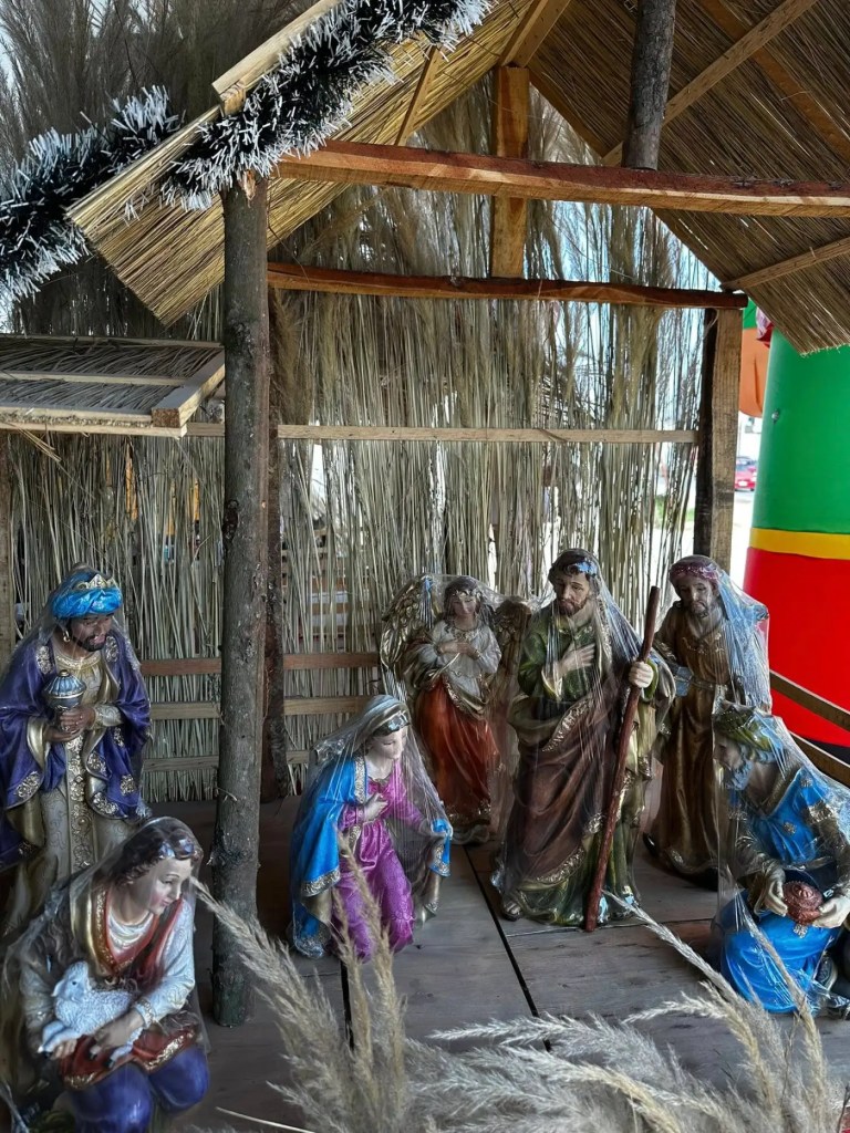 Un nacimiento navideño con figuras de resina y un techo de palma, decorado con ramas secas y un fondo colorido.