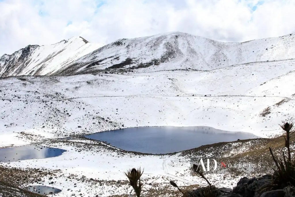 Vista panorámica del Nevado de Toluca cubierto de nieve, con un lago reflejando el paisaje montañoso en el fondo.