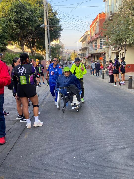 Un grupo de corredores y participantes se preparan para la carrera atlética, incluyendo a un atleta en silla de ruedas, en un entorno urbano.