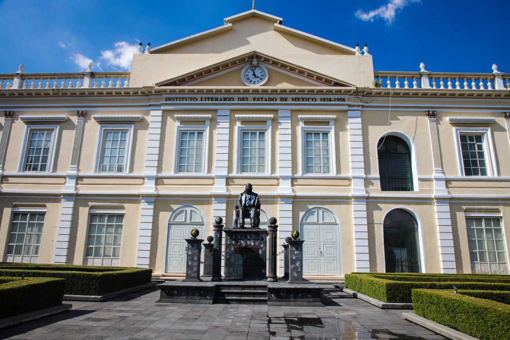 Facade of the Instituto Literario del Estado de Mexico, featuring a statue in front, with a blue sky and decorative landscaping.