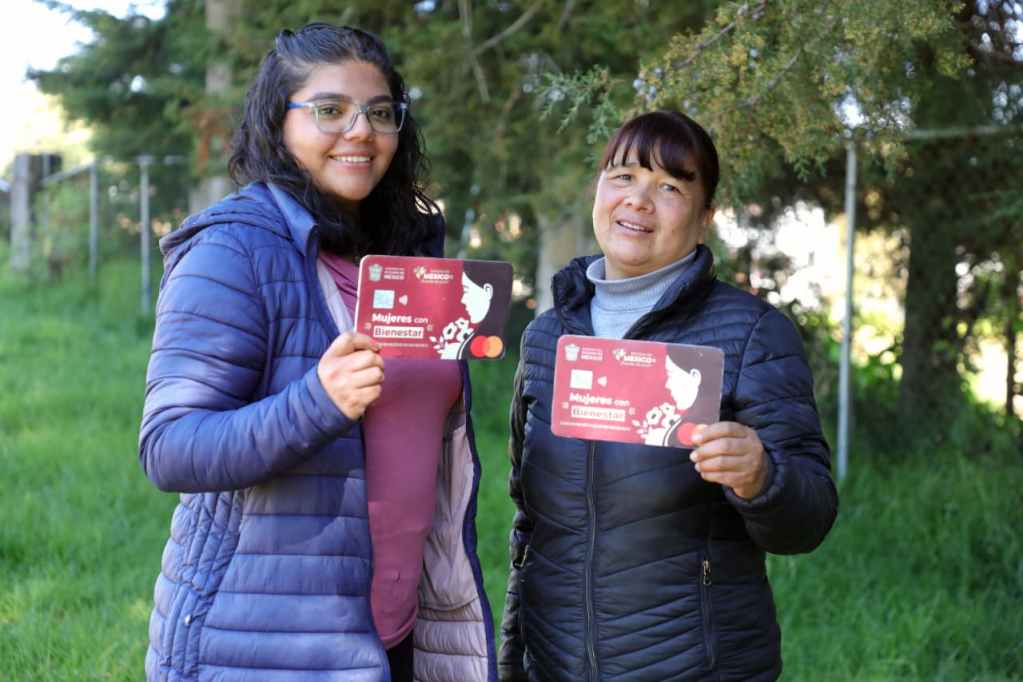 Dos mujeres sosteniendo tarjetas del programa 'Mujeres con Bienestar' en un área verde.