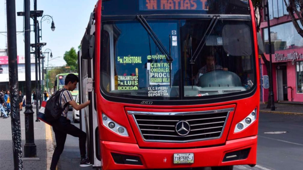 Un joven está subiendo a un autobús rojo de transporte público en una calle concurrida, con señalización visible en el frontal del vehículo.