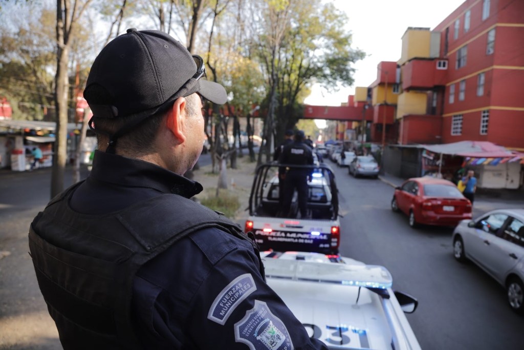 Policía municipal en patrullaje por calles de un barrio residencial, con vehículos de seguridad en segundo plano.