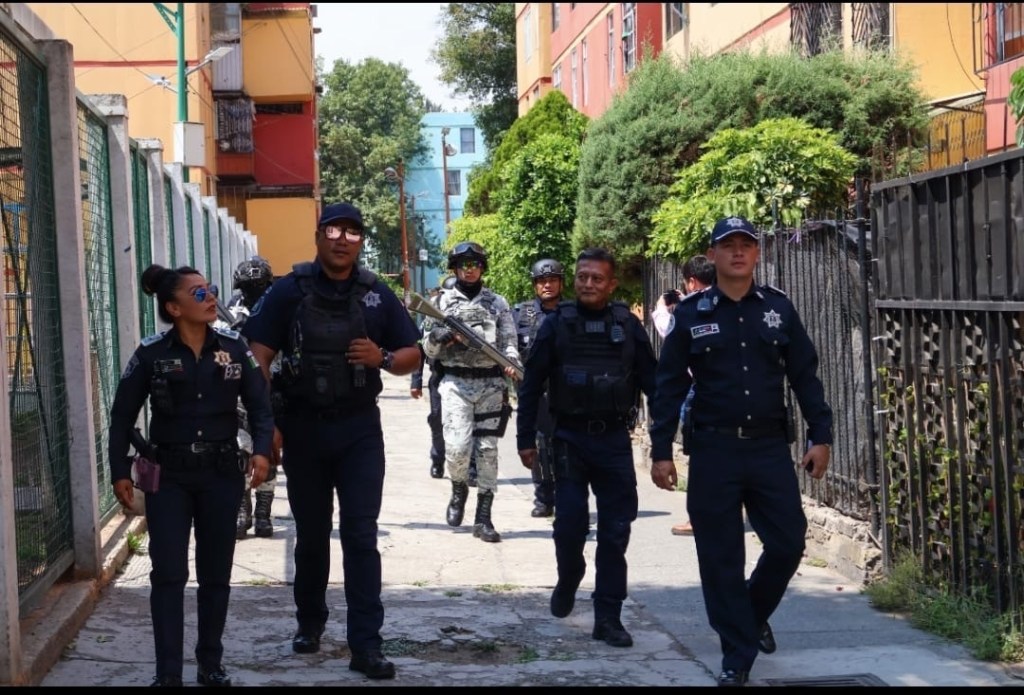 Grupo de policías caminando por una calle residencial, algunos con equipamiento táctico y otros en uniforme, mientras realizan labores de patrullaje y vigilancia.