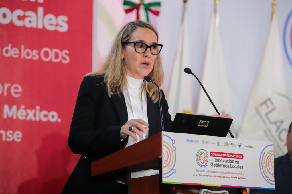 Una mujer con cabello largo y rubio, usando un saco negro y blusa blanca, habla en un podio durante el Seminario Internacional de Gobiernos Locales en San Mateo Atenco. En el fondo, se pueden ver banderas y un cartel del evento destacando la innovación en gobiernos locales y los ODS.