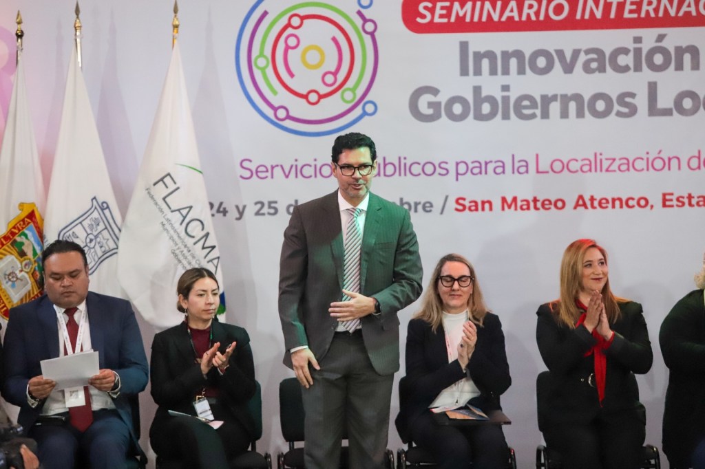Un grupo de personas en un seminario internacional sobre gobiernos locales, con un speaker de pie frente a una presentación, mientras los demás asisten en un ambiente formal.