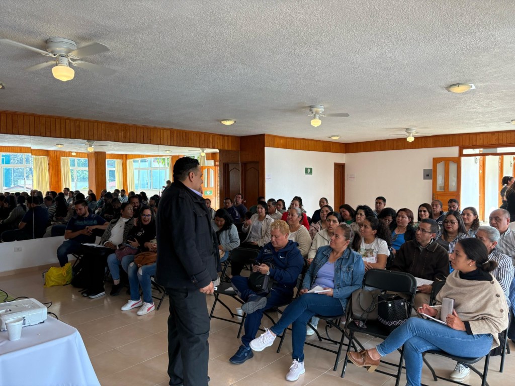 Un grupo de personas participan en una sesión de capacitación, mientras un orador se dirige a ellos. Se observa un ambiente de aprendizaje con una disposición de asientos en un salón bien iluminado.